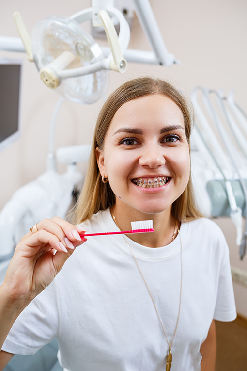 A young woman in a white t-shirt sits in a dental office, smiles and holds a toothbrush in her hands. Girl with braces shows oral care. Dentistry, dental treatment pokaz koron zębowych
